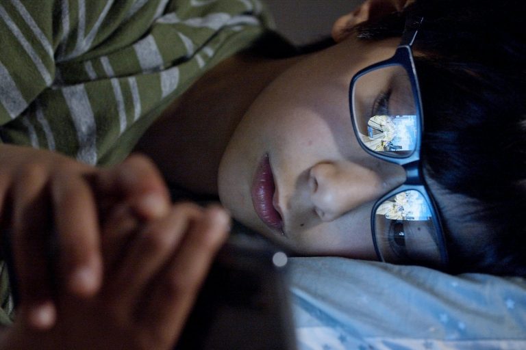 A child lying in bed at night using a smartphone, with screen reflection on eyeglasses.